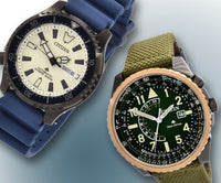 A white dial blue strap watch next to a green dial and strap watch. They are on a light blue and white backgroud. ,