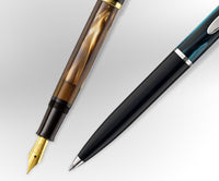 A Pelikan fountain pen with a gold nib and gold tone body next to a Pelikan ballpoint pen with a marbled black body on a light silver and white background. ,
